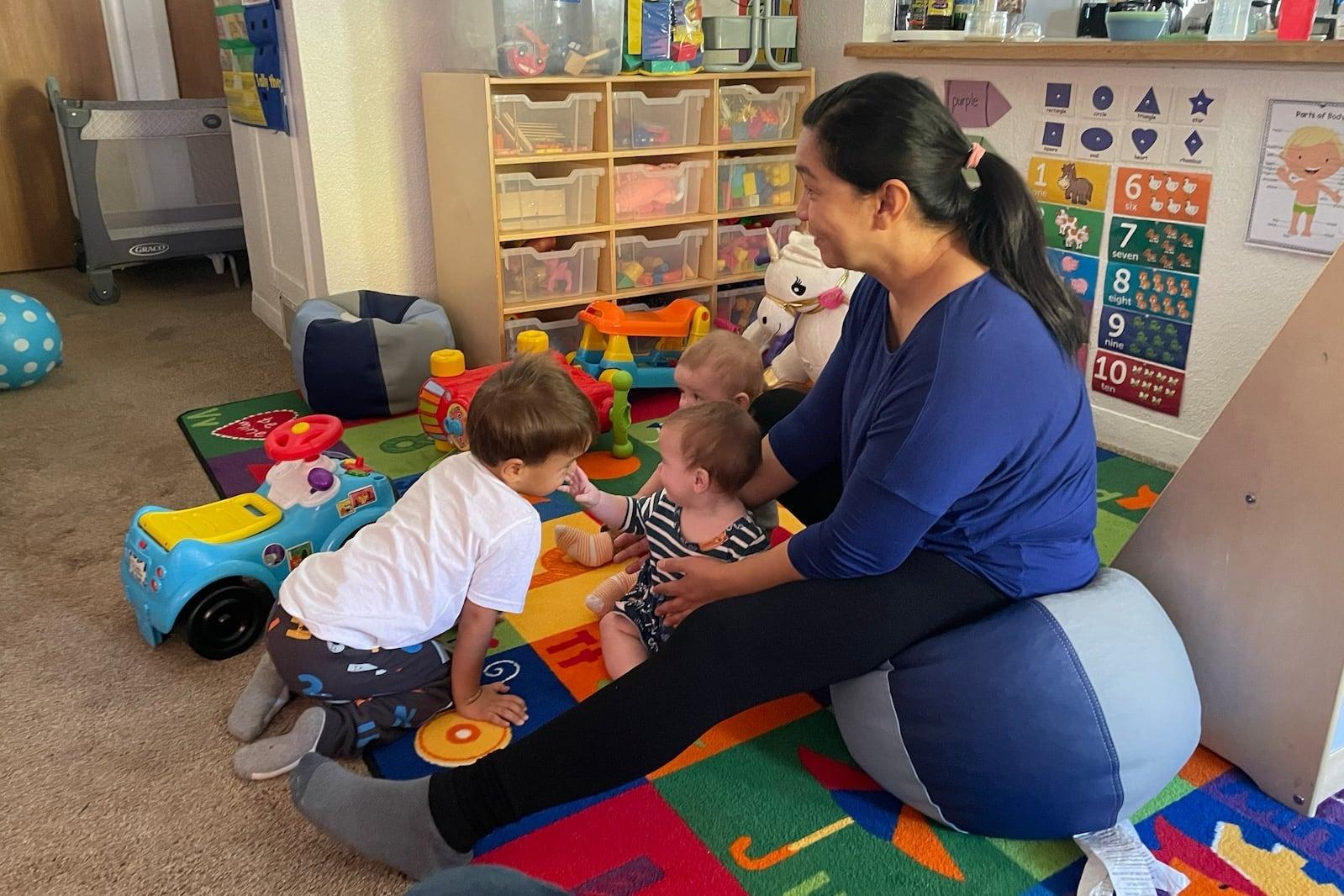 A woman with black hair in a ponytail blue shirt leggings sits on a round bean bag with children surrounding her