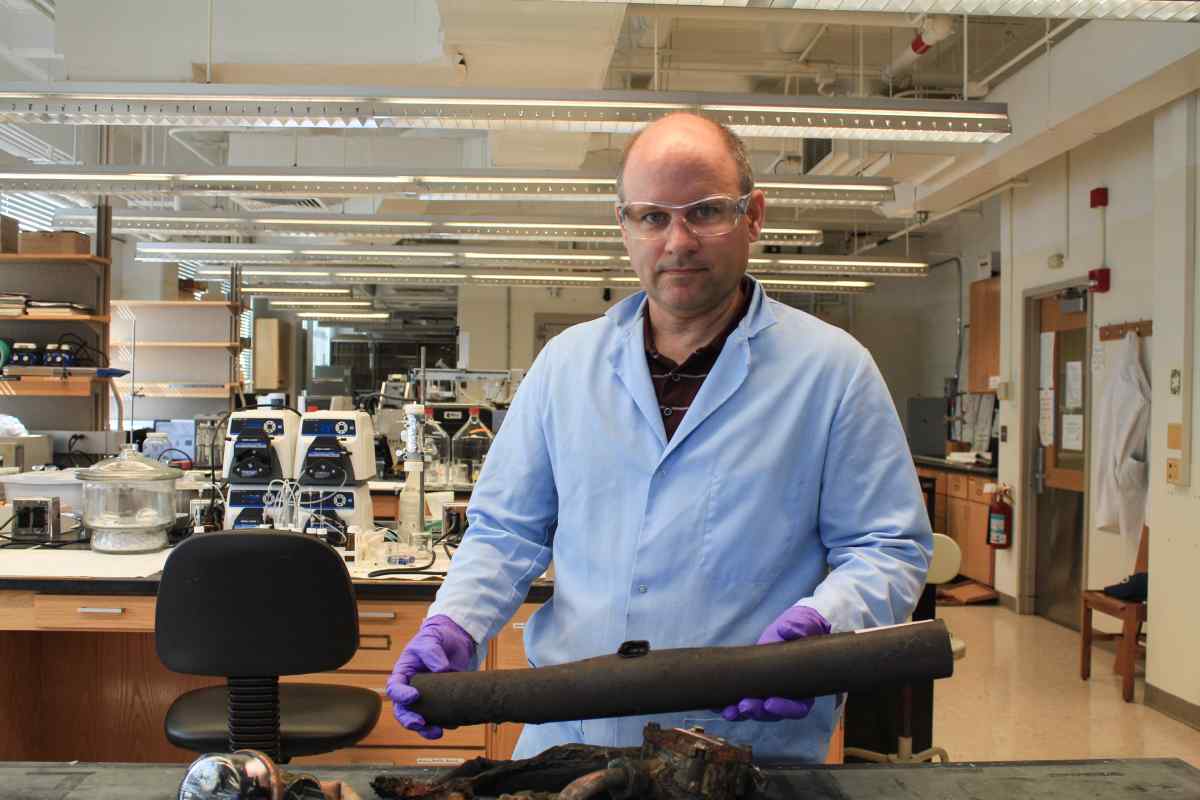 A man in a blue lab coat goggles and purple gloves holding a charred item in a lab