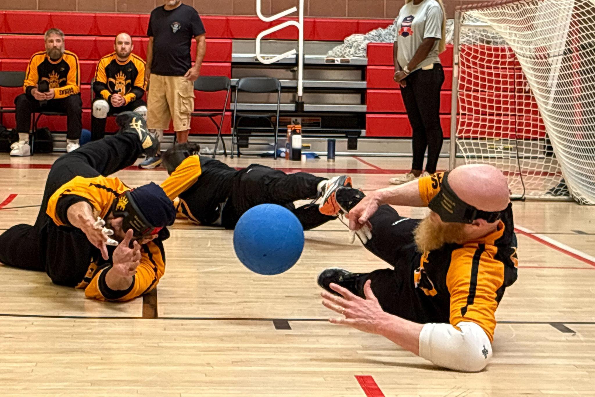 Two people dive for a blue ball on the floor with yellow and black jerseys on.