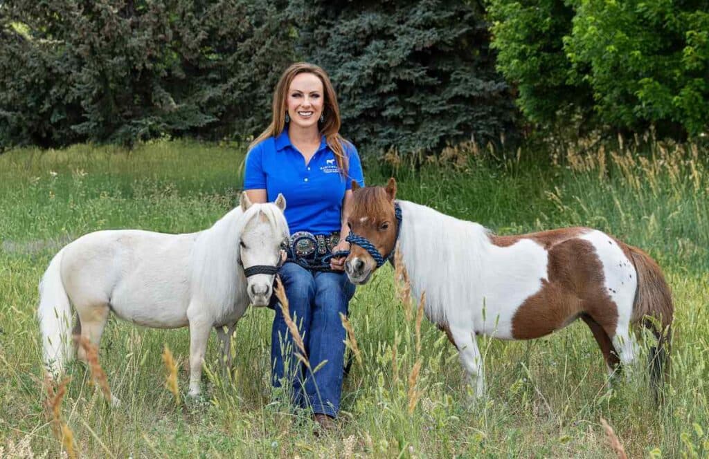A woman sits in a field between two mini horses on either side of her facing the camera