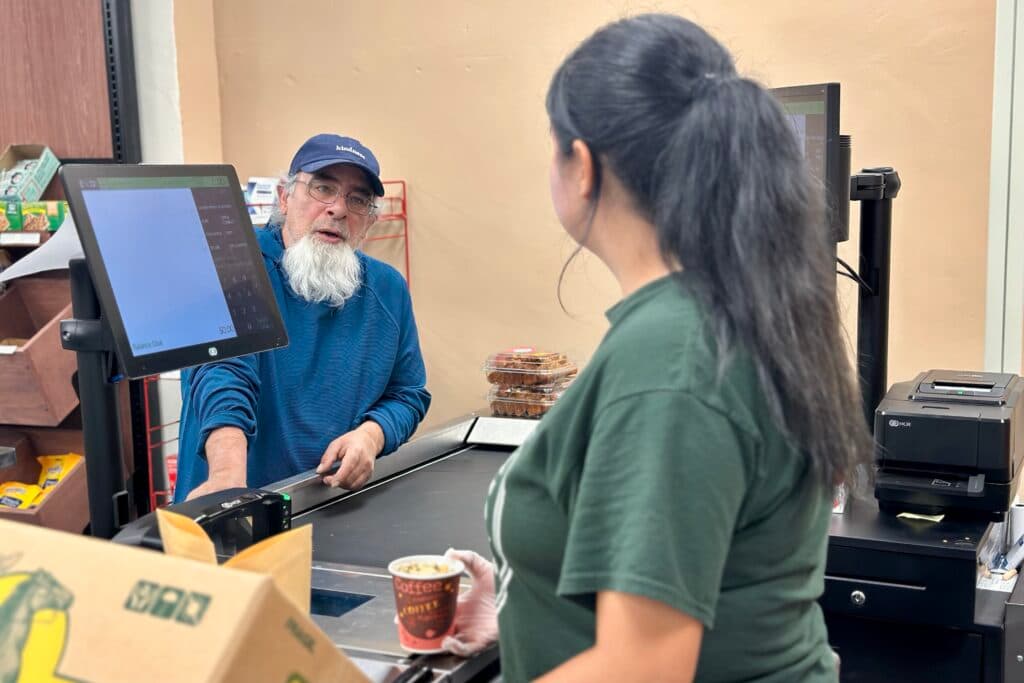 A man with a gray goatee and blue baseball cap speaks with a market teller.