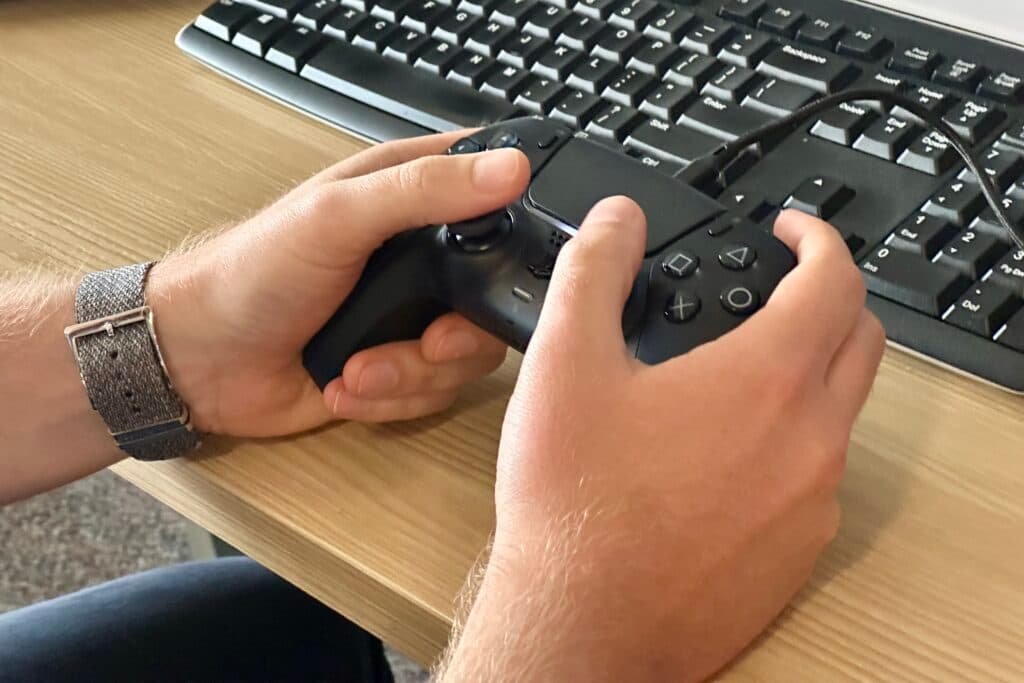 This a closeup of a person's hands holding a Playstation 5 controller in front of a keyhboard