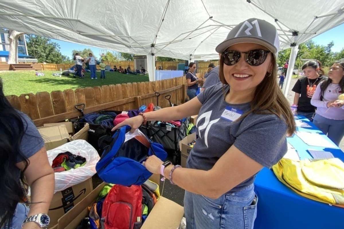 Woman in blue jeans and dark blue t-shirt sorts through multicolored backpacks while looking at the camera.