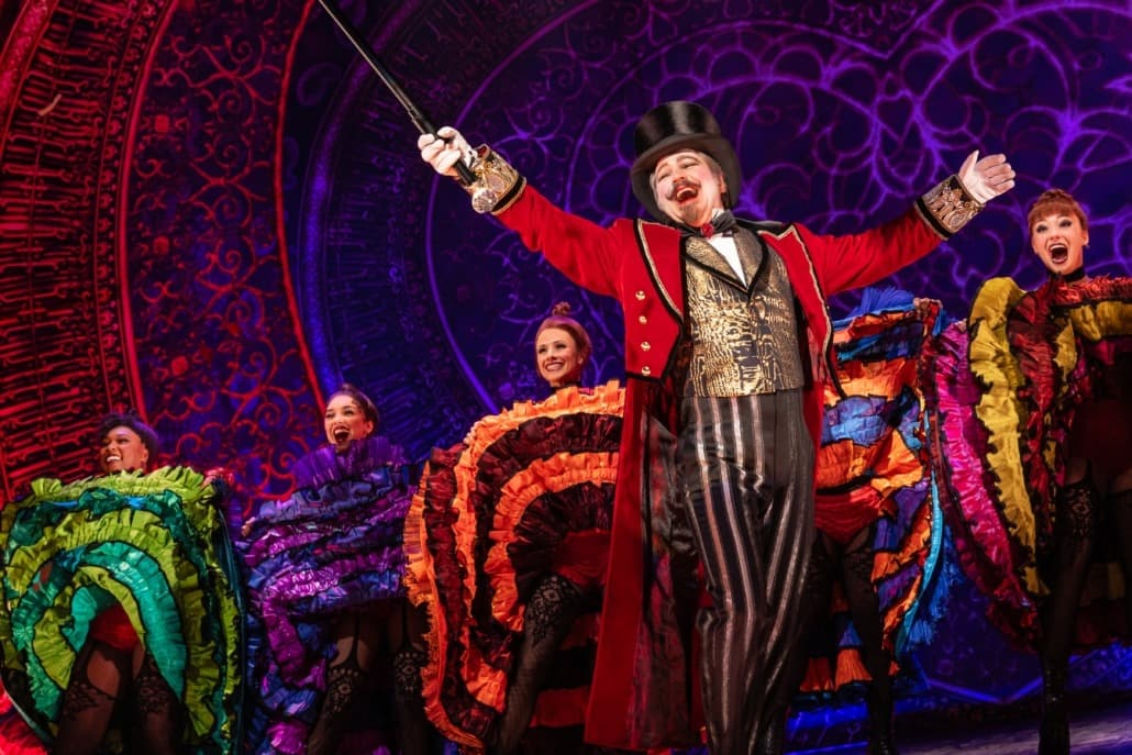 A man in a top hat and extravagant outfit raises his cane in the air as can-can dancers kick behind him.