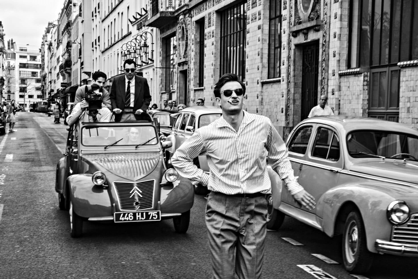 A black-and-white shot of a man walking down the street, smoking a cigarette, with a car ful of men behind him, one filming his back as he walks.