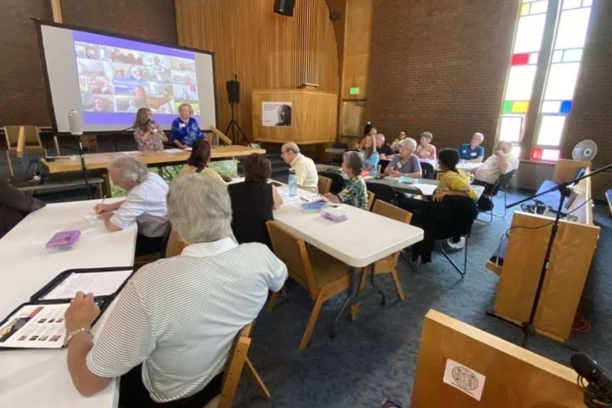 A group of people are gathered around tables, looking at slides projected on a screen in front of a church. They are discussing a ballot initiative to create single payer health insurance.
