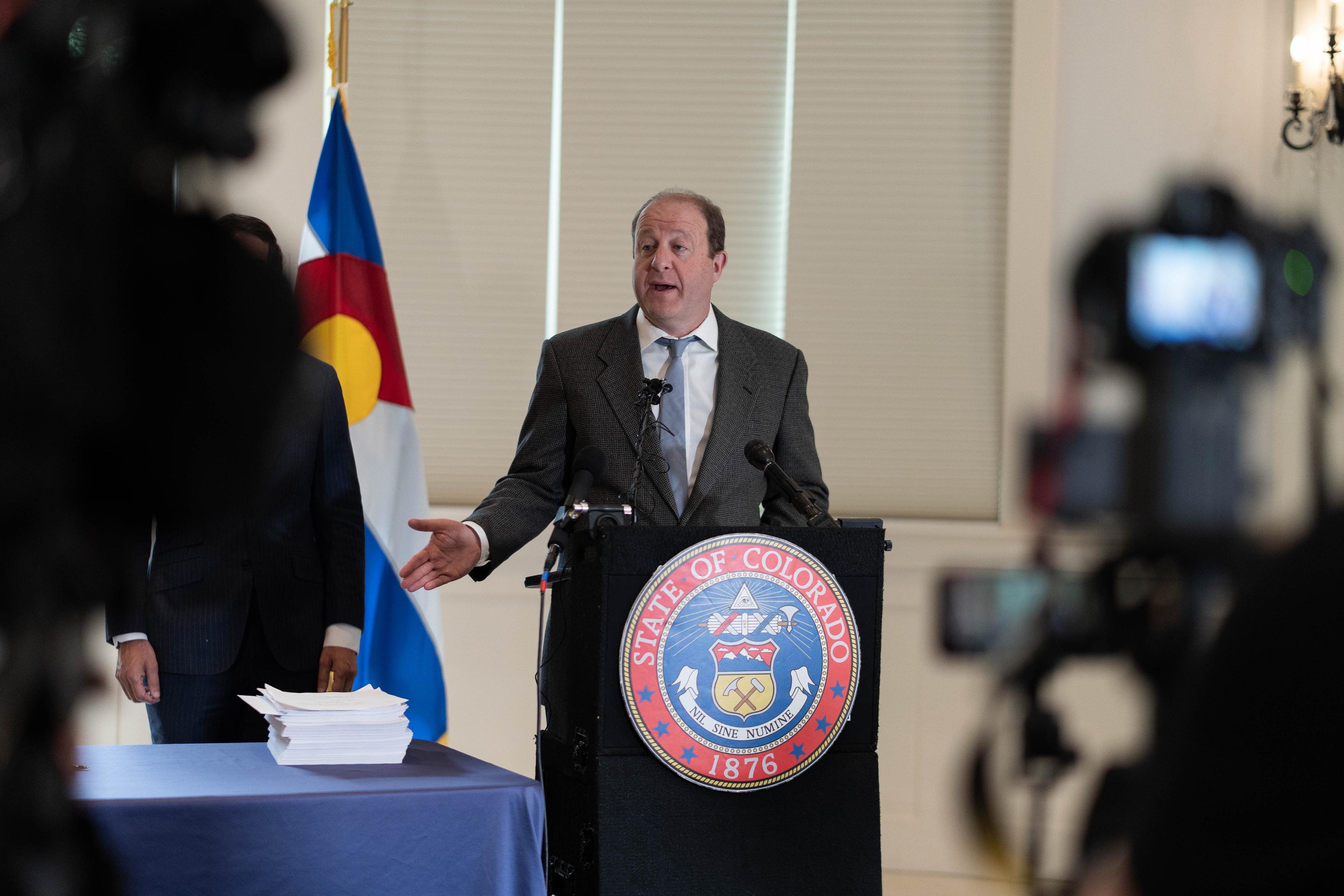 Gov Jared Polis stands at a lectern, with a Colorado flag behind him. He's speaking and gesturing.