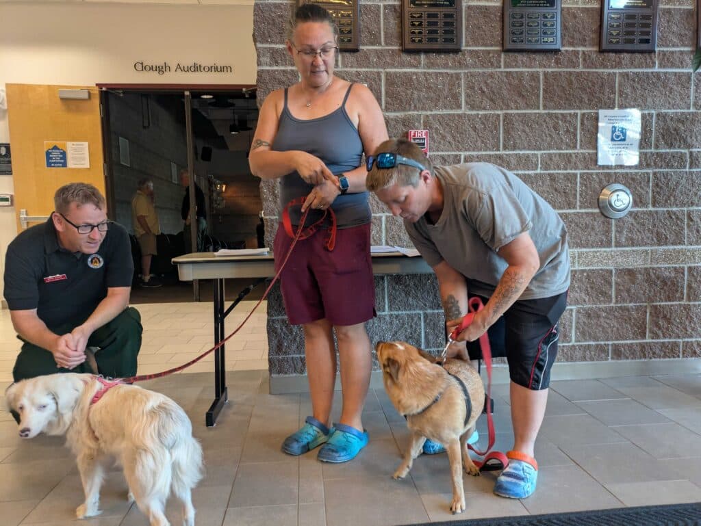 Two people stand with their dogs on leashes inside a building.