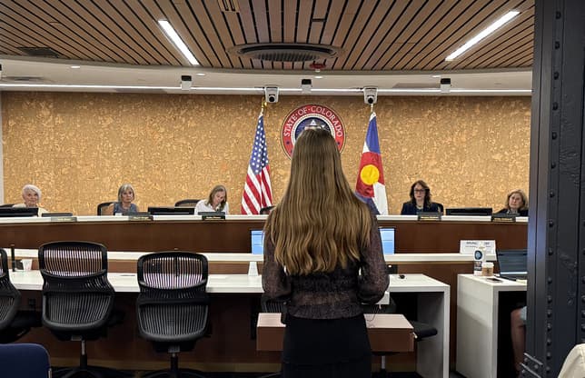 A girl stands with her back to the camera as she faces a panel of people in a boardroom. There is a seal that says State of Colorado on the wall behind the board members along with a U.S. flag and a Colorado flay on poles.