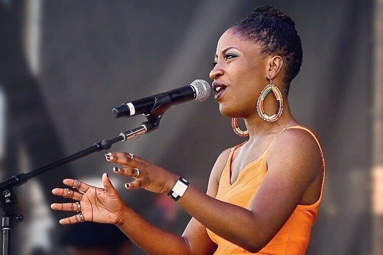 A woman wearing an orange dress is seen sideways singing into a microphone looking to the left.