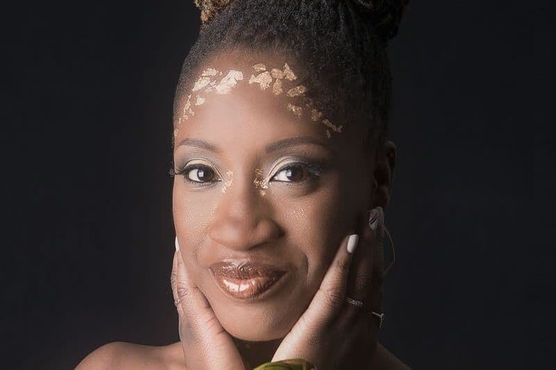 Photo shows a Black woman with her hands against her cheeks with her hair up and she is smiling.