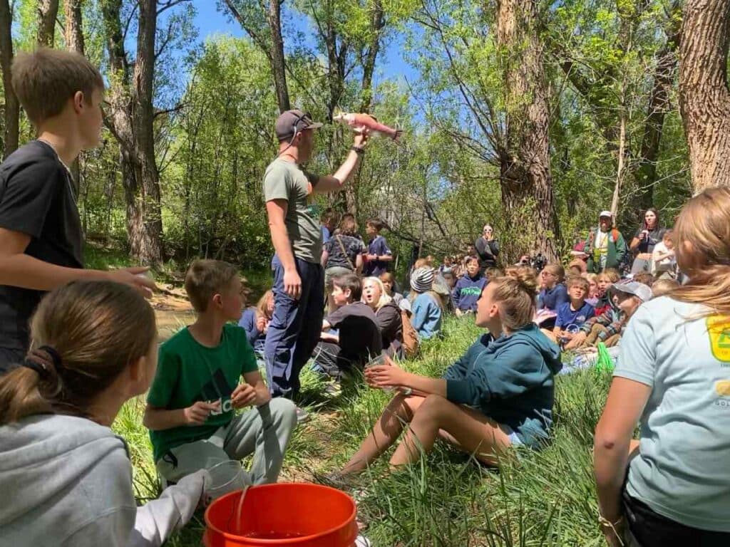 A group of students sit in in the grass among trees looking up at a teacher
