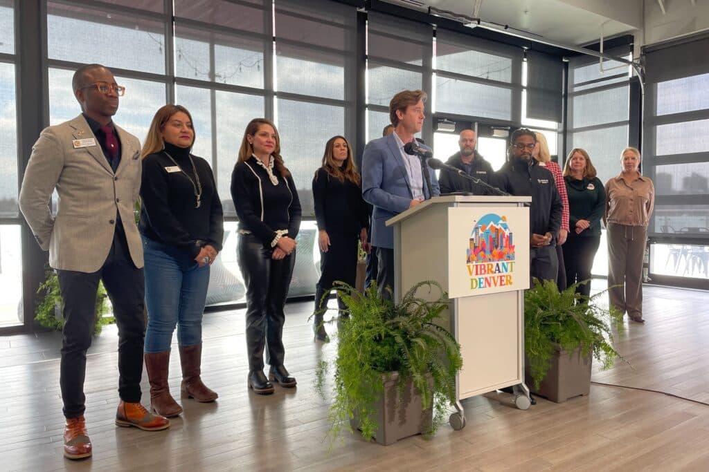 Denver Mayor Mike Johnston at a podium with 10 people standing behind him