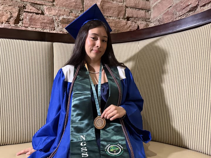 A Latina sits in her blue graduation regalia that includes a green sash that says NGSS and a medallion that certifies her as climate literate.