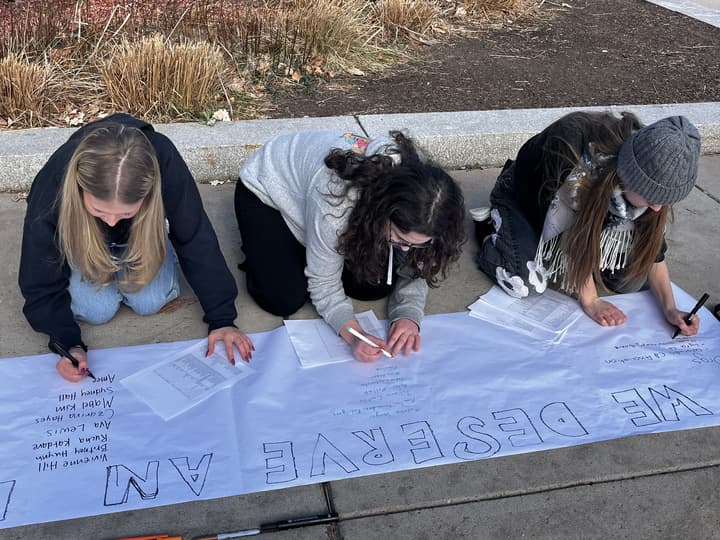 Three young women kneel on the sidewalk as they write messages on a banner.