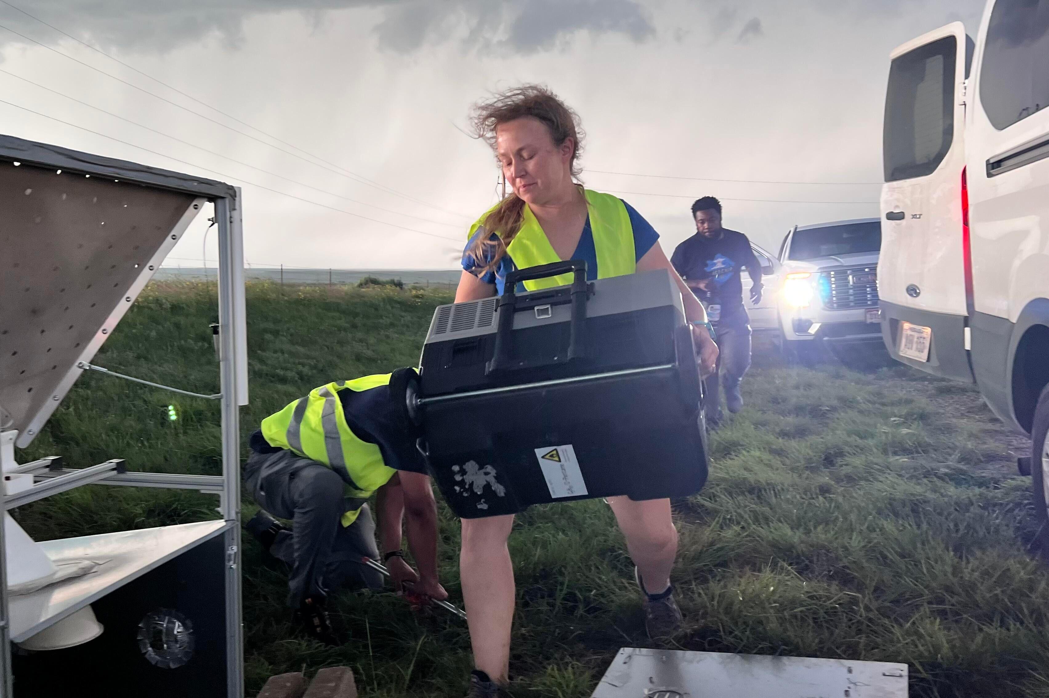 A person in a bright yellow safety vest carries a large black box outside several parked cars in a large field with storm clouds above.