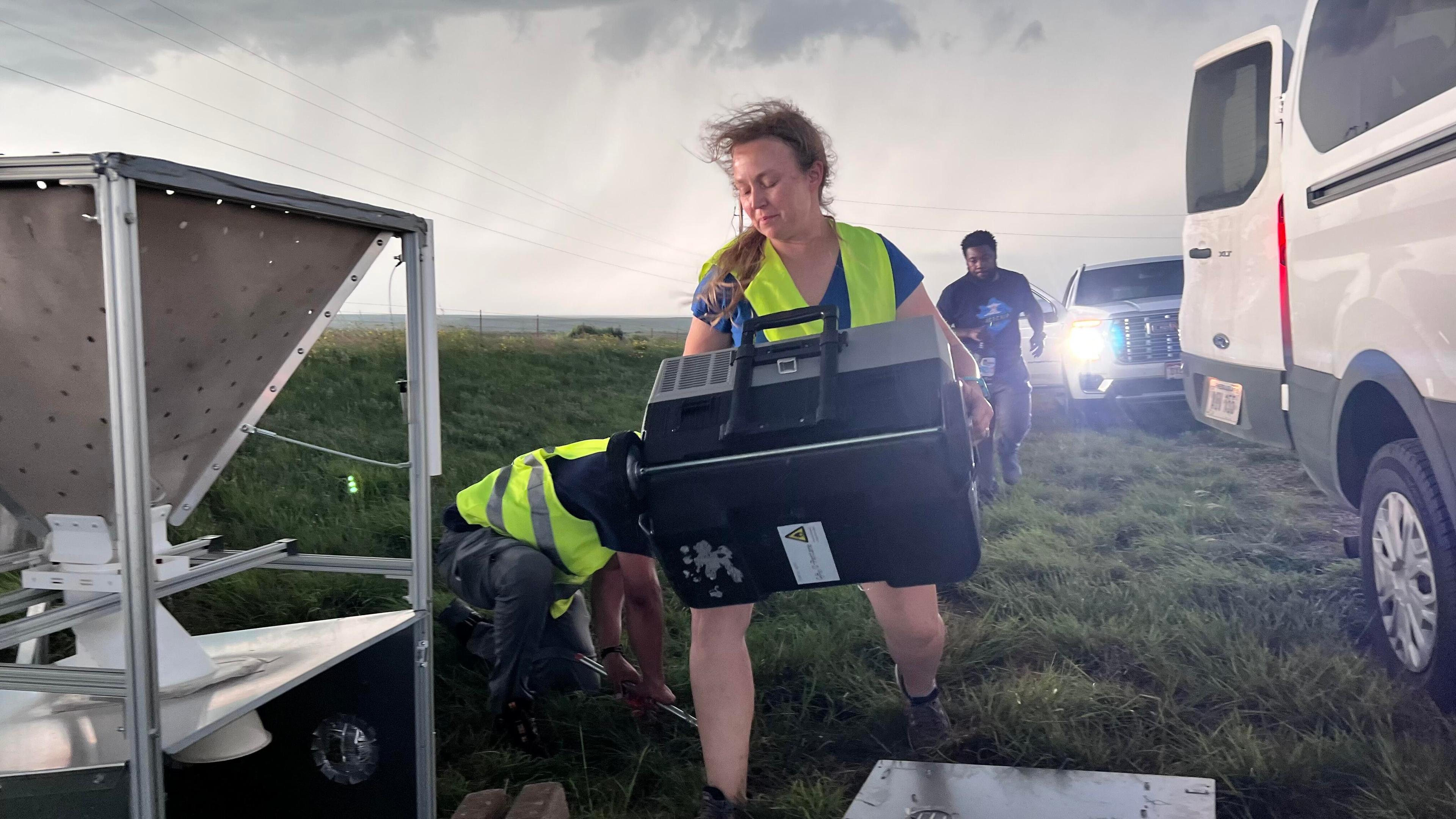 A person in a bright yellow safety vest carries a large black box outside several parked cars in a large field with storm clouds above.