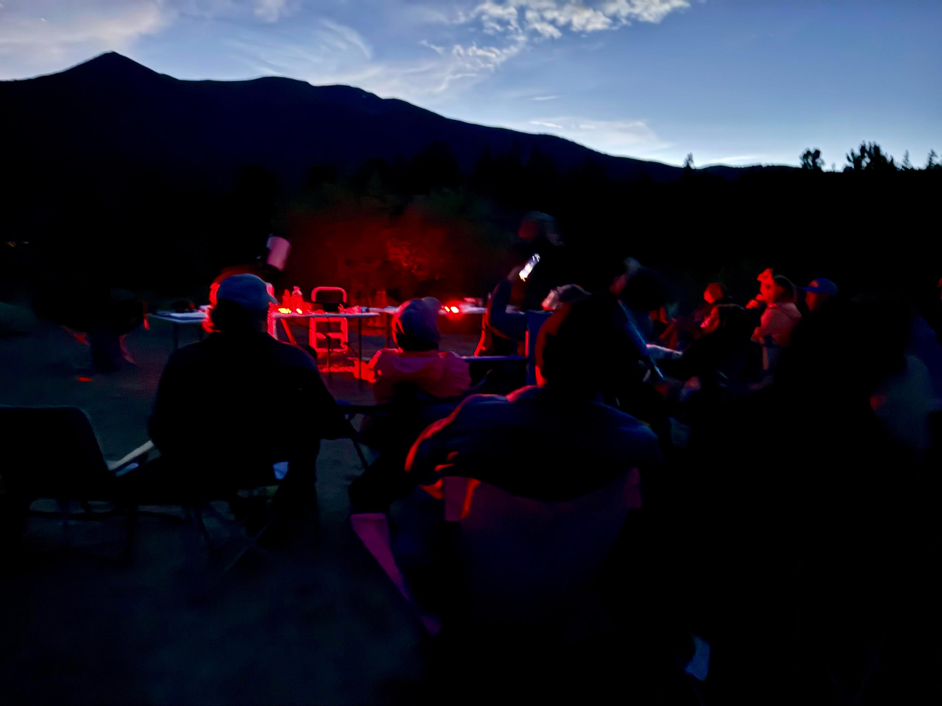 A group of people is seated outside near the mountains at night. Only parts of them are illuminated by glow from red lights.