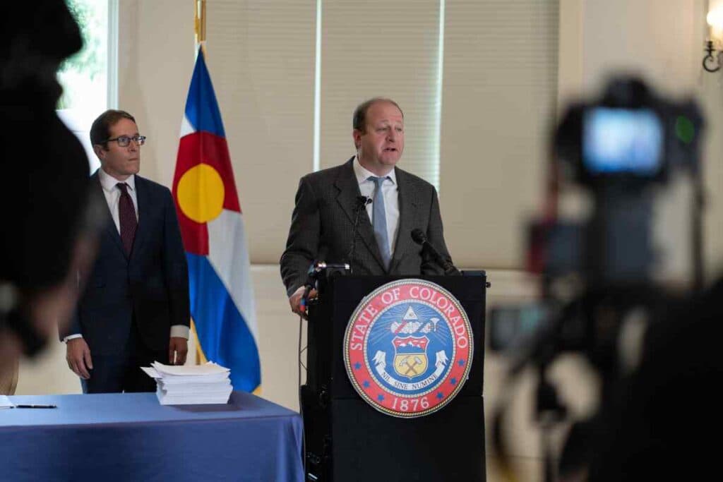 Jared Polis in a grey suit blue tie at a podium speaking into a mic with a man in glasses and a table and flag behind him