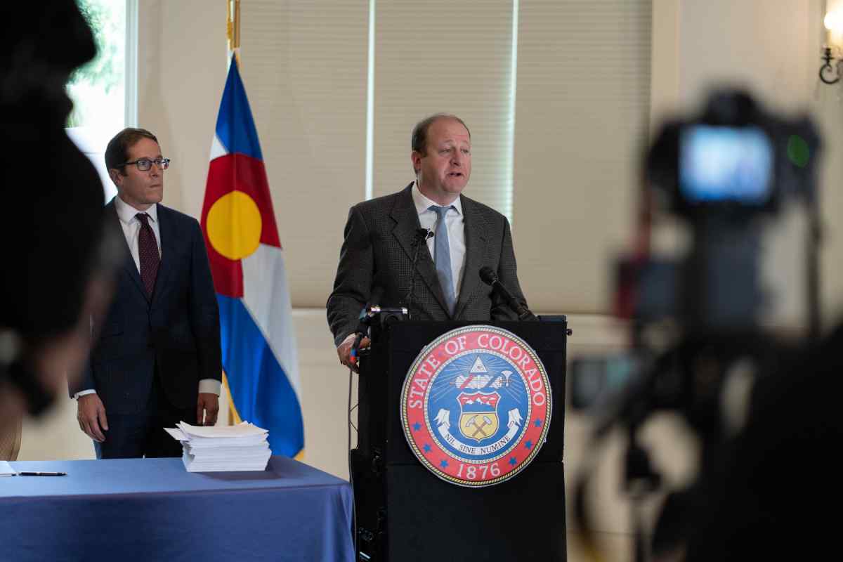 Jared Polis in a grey suit blue tie at a podium speaking into a mic with a man in glasses and a table and flag behind him