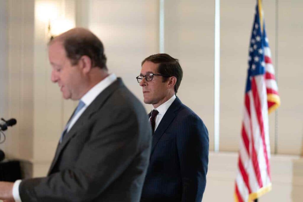 The camera is focused on a man in a navy suit with glasses in the foreground blury is a man in a grey suit the American flag is blurry in the background