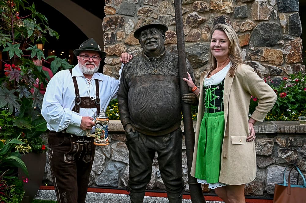 Image shows a man in traditional lederhosen holding a stein to the left of a bronze sculpture of a man named Helmut Fricker. On the right is a woman in a green dress with a beige coat. They are in front of a stone pillar with plants on left side of the photo.