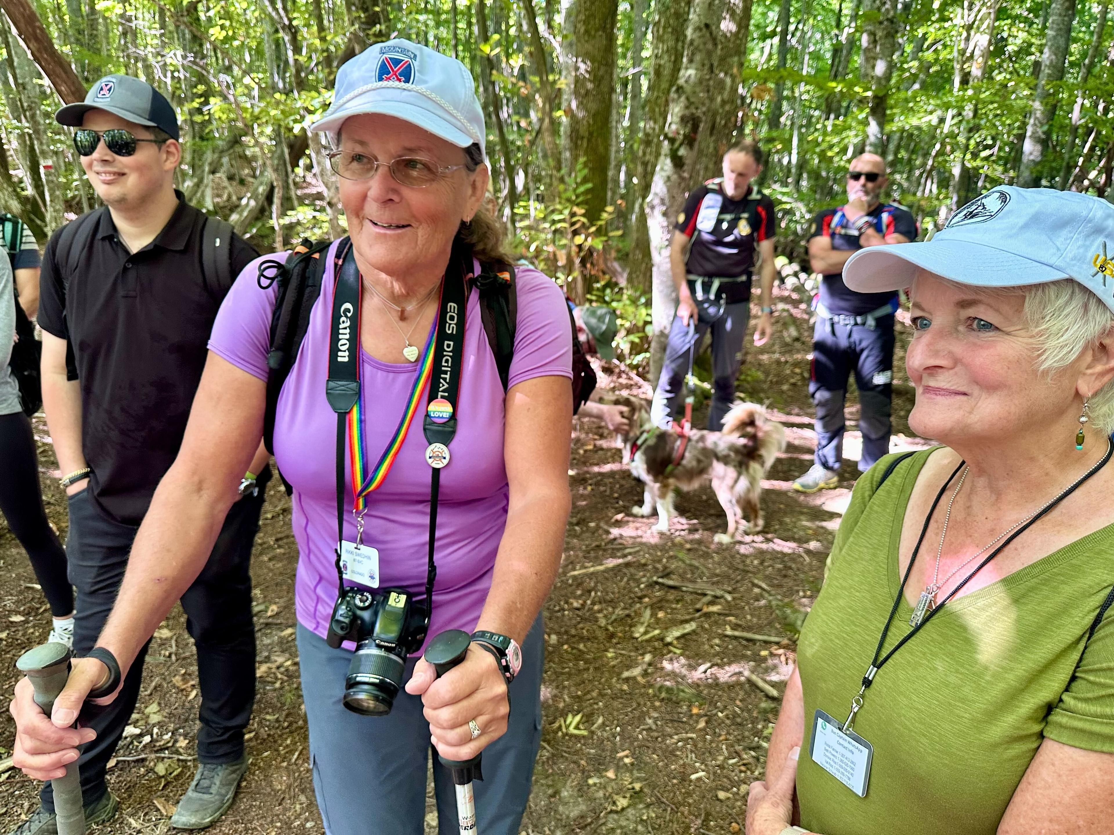 A woman in a purple short and baseball cap smiles as she stands with hiking poles next to other people in a wooded area on an Italian mountain.