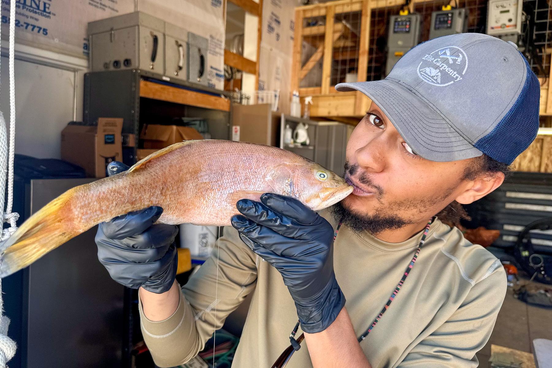 A man in a ballcap makes a kissy face at a brown fish he’s holding.