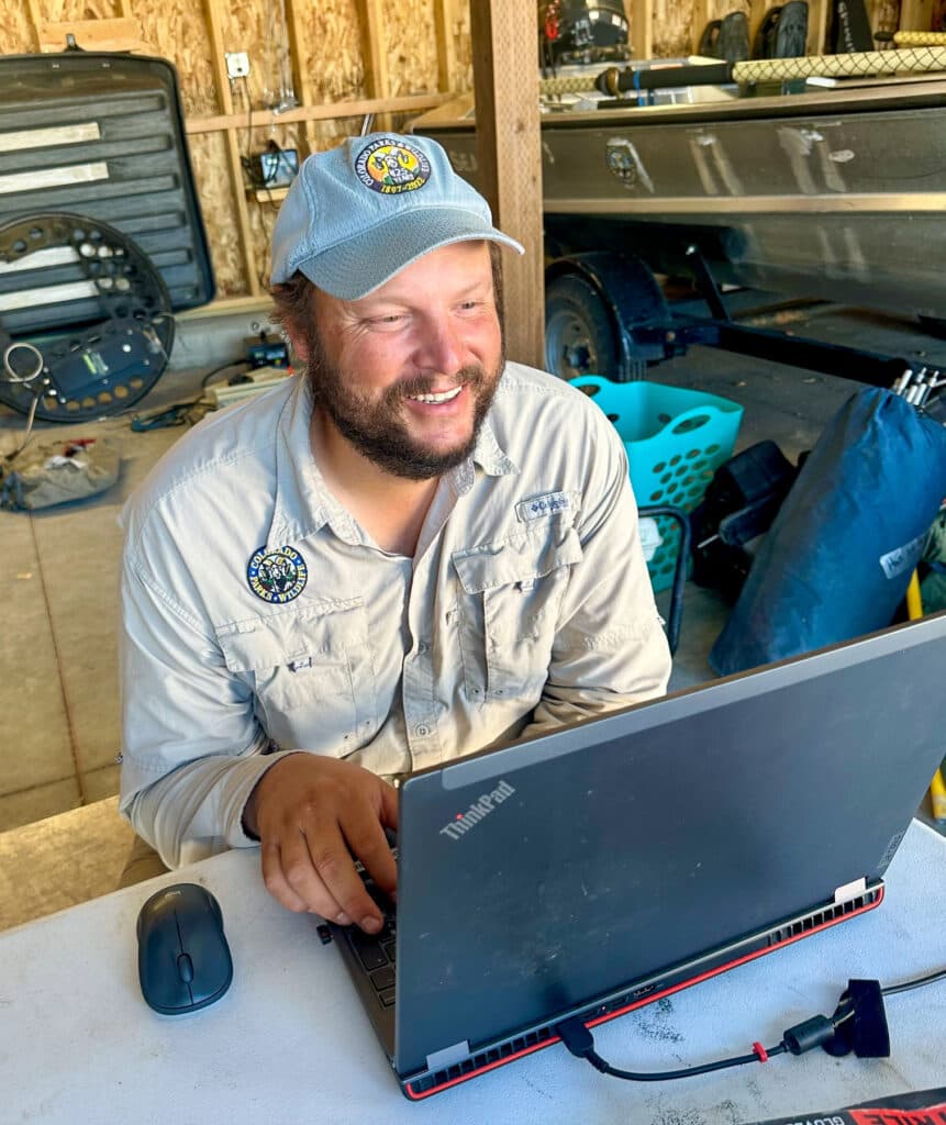 A smiling man in a ballcap sits typing on a laptop. A boat is in the background to his left.