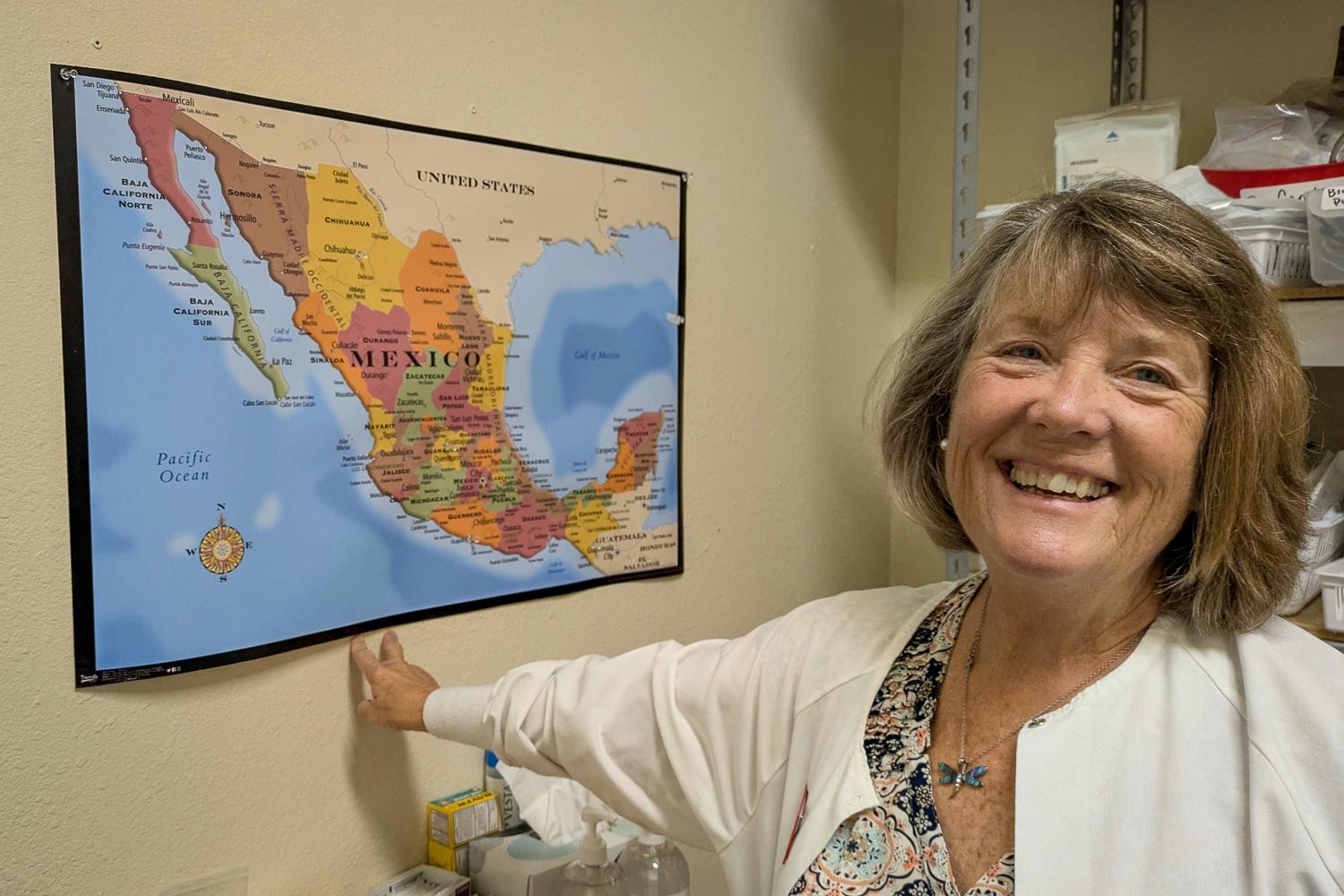 A colorful map of Mexico is framed on the wall. To the right, a woman with short hair is smiling as she points to the map.