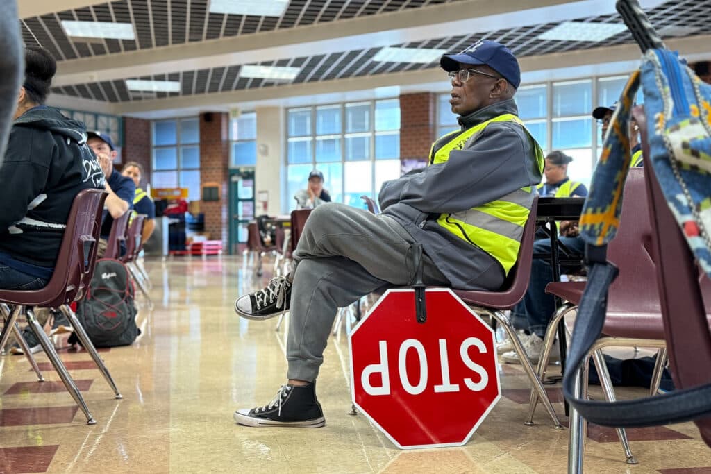 A man wearing a crossing guard hat and yellow vest sits with his big stop sign resting next to his chair.