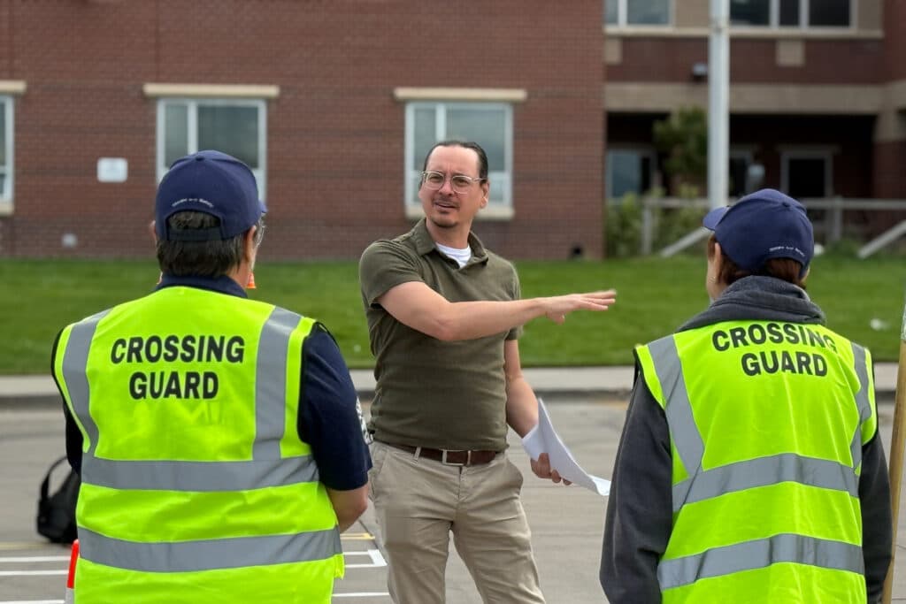 In the foreground, the backs of two people wearing yellow reflective vests that read Crossing Guard. The two are looking at a man in glasses in khakis and a green polo shirt gesturing to his left.