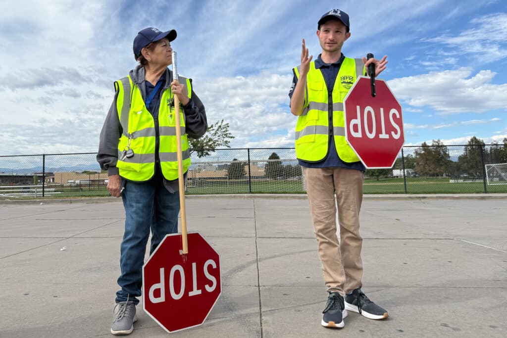 A woman, left, looks over at a man as he speaks in a parking lot. He is holding his hands out in a gesture as he hold a stop sign with one hand. The woman has a stop sign on a longer pole, which she rests on the ground. Both are wearing crossing guard hats and yellow reflective vests.
