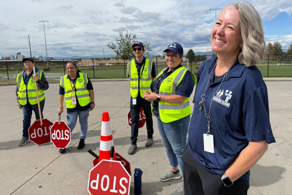 A group of five people stand outside in a parking lot behind a traffic cone. They all have big red stop signs on handles. Four of the five people are wearing yellow reflective vests.