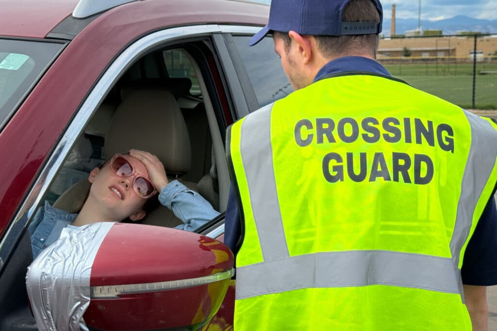 A woman in the driver's seat of a car leans bacl and puts her left hand on her forehead. A man in a crossing guard vest looks at the woman. His back is to the camera.