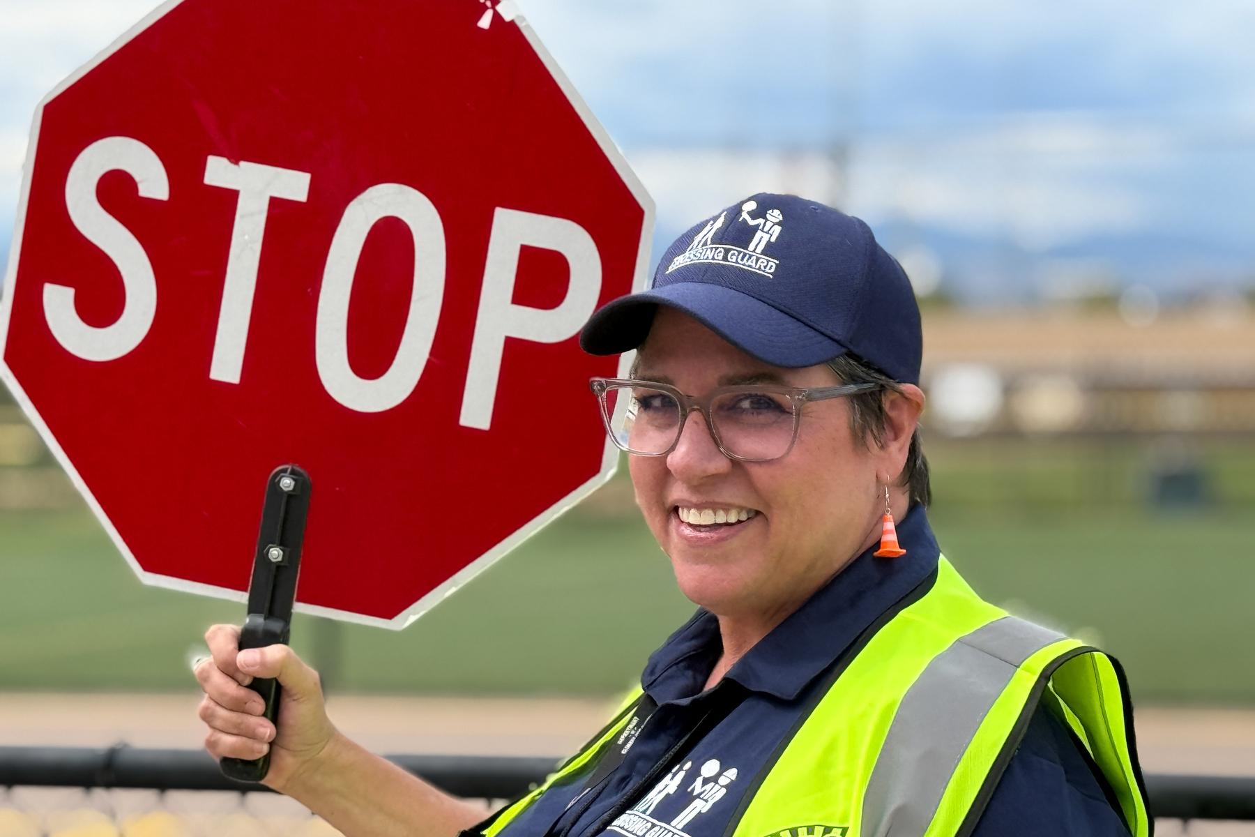 A woman in glasses and a navy blue hat that reads "Crossing Guard" holds up a big red stop signs as she looks at the camera. She is wearing earrings that are little traffic cones and the yellow reflective vest of her job.