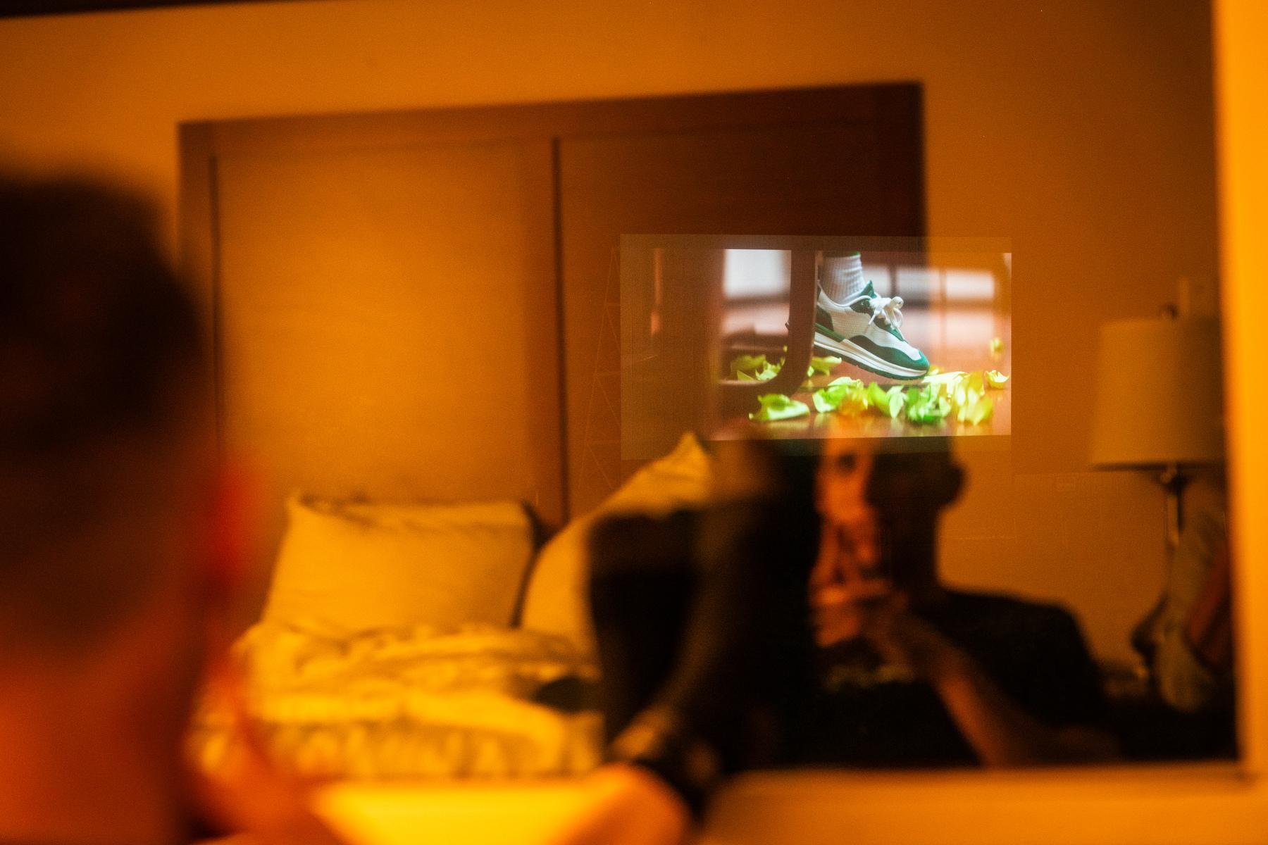 A person is sitting in a bedroom, watching a reflection of a TV screen in a mirror. The TV is displaying an image of sneakers. The man is holding a remote control in his hand, likely controlling the TV. The room is dimly lit.