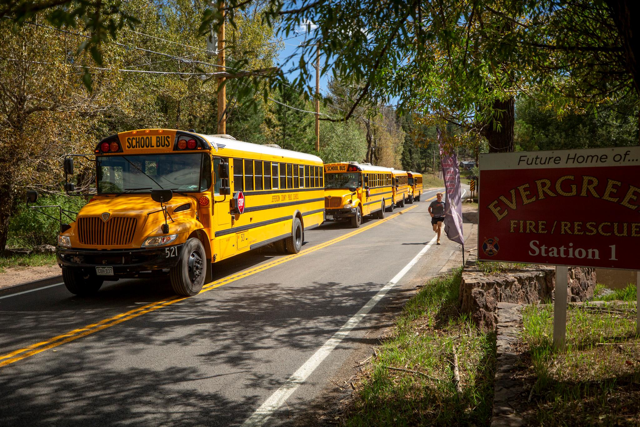 Three yellow school buses are parked along a tree-lined road while a person jogs past on the shoulder. A red sign on the right reads “Future Home of ... Evergreen Fire/Rescue Station 1.”