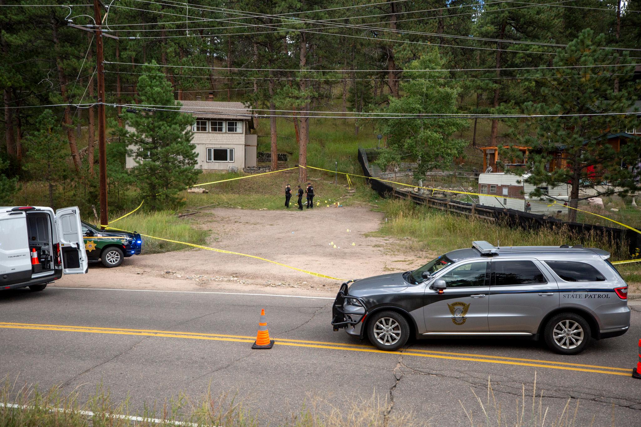 A state patrol SUV and a white van block a road, with yellow tape marking off a dirt area in front of a structure. Three officers stand inside the taped area, surrounded by trees and utility wires.