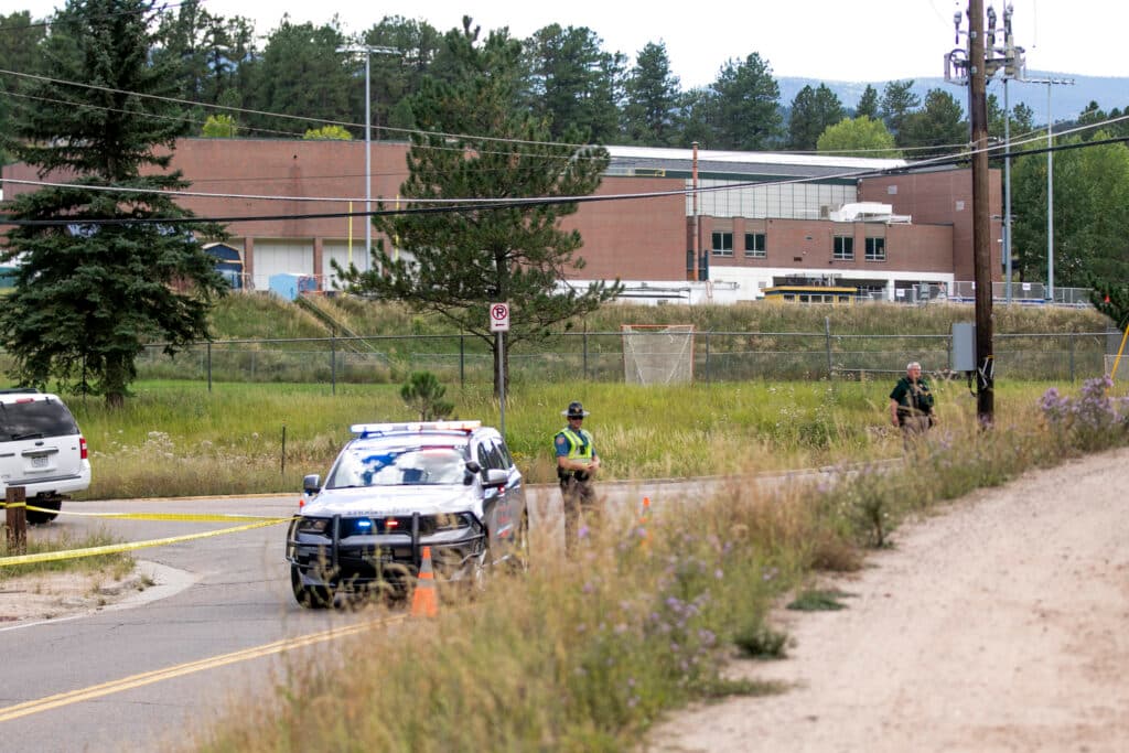 A police car with flashing lights, two officers, and yellow tape block a rural road near a brick building surrounded by trees and grassy areas.