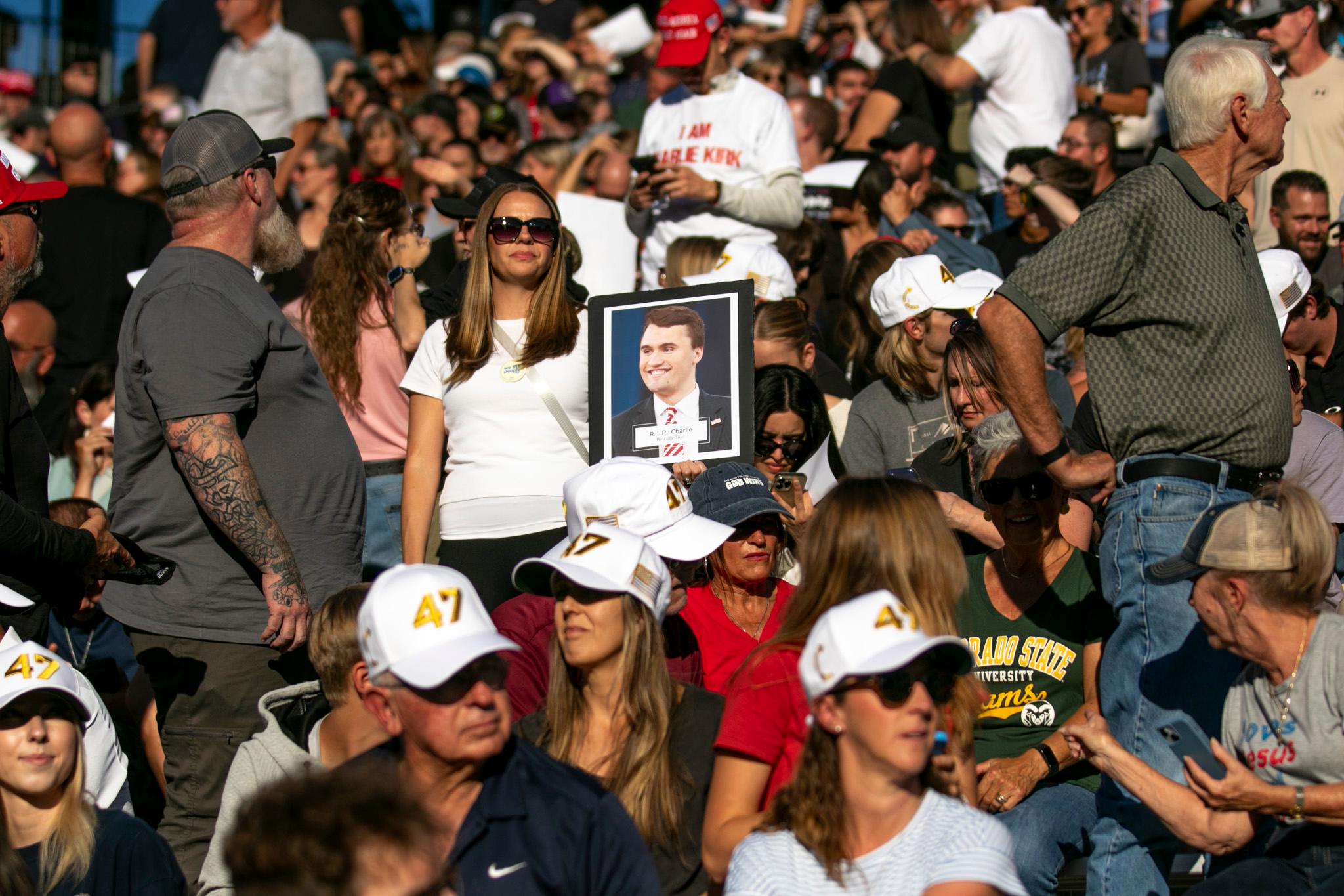 A crowd sits outdoors wearing hats, many in white with the number 47. One woman stands holding a portrait of a smiling man in a suit. Some people wear sunglasses and shirts with various logos. A woman holds a photo of Charlie Kirk.