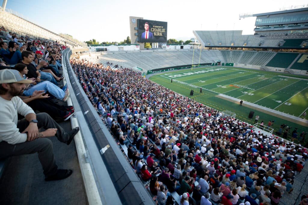 The green grass of a football stadium is surrounded by bleachers; one section, closest to the camera, is filled with thousands of people. A megatron in the background reads "in memory of Charlie Kirk."