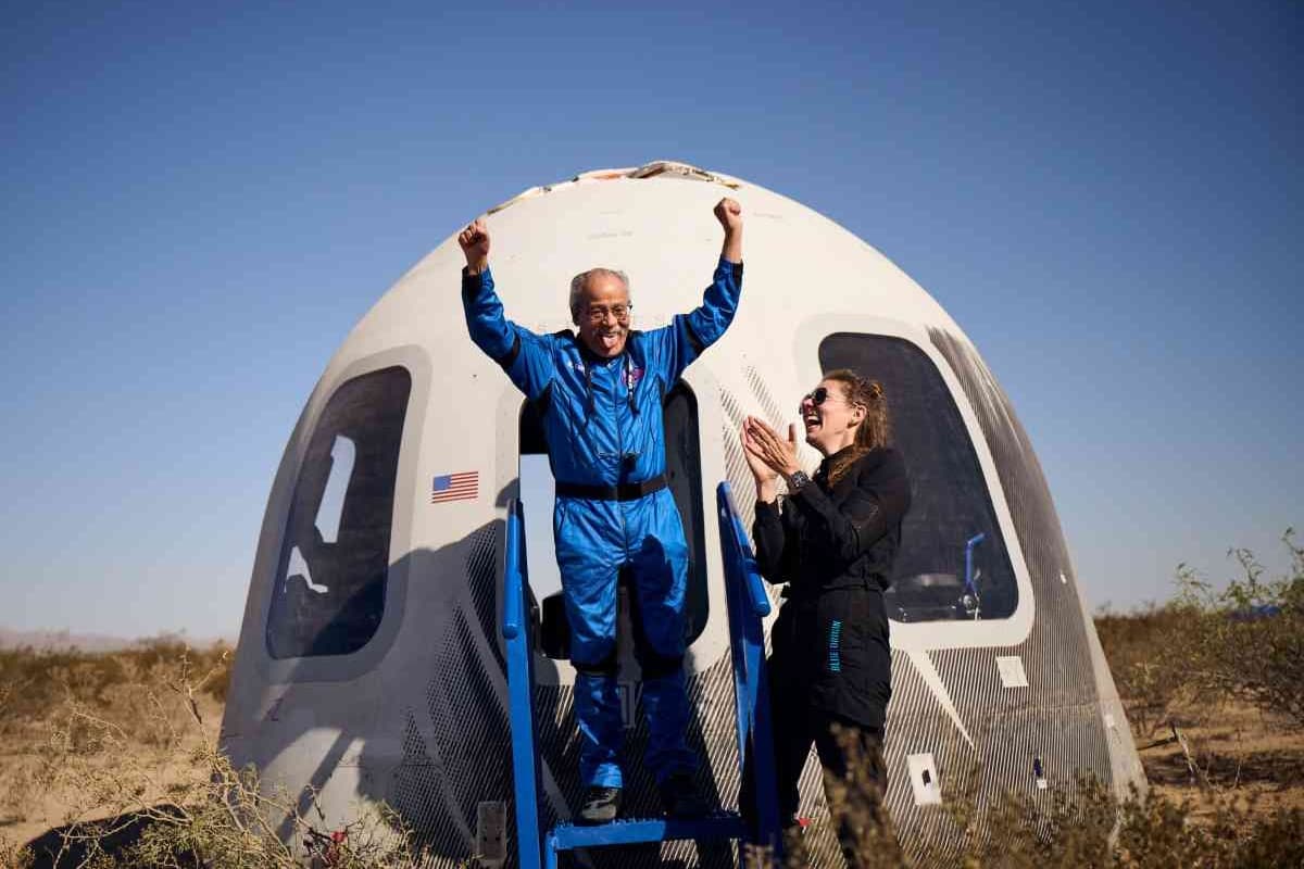 A man in a blue astronaut uniform cheers with both hands up a woman in a black jumpsuit laughs and claps