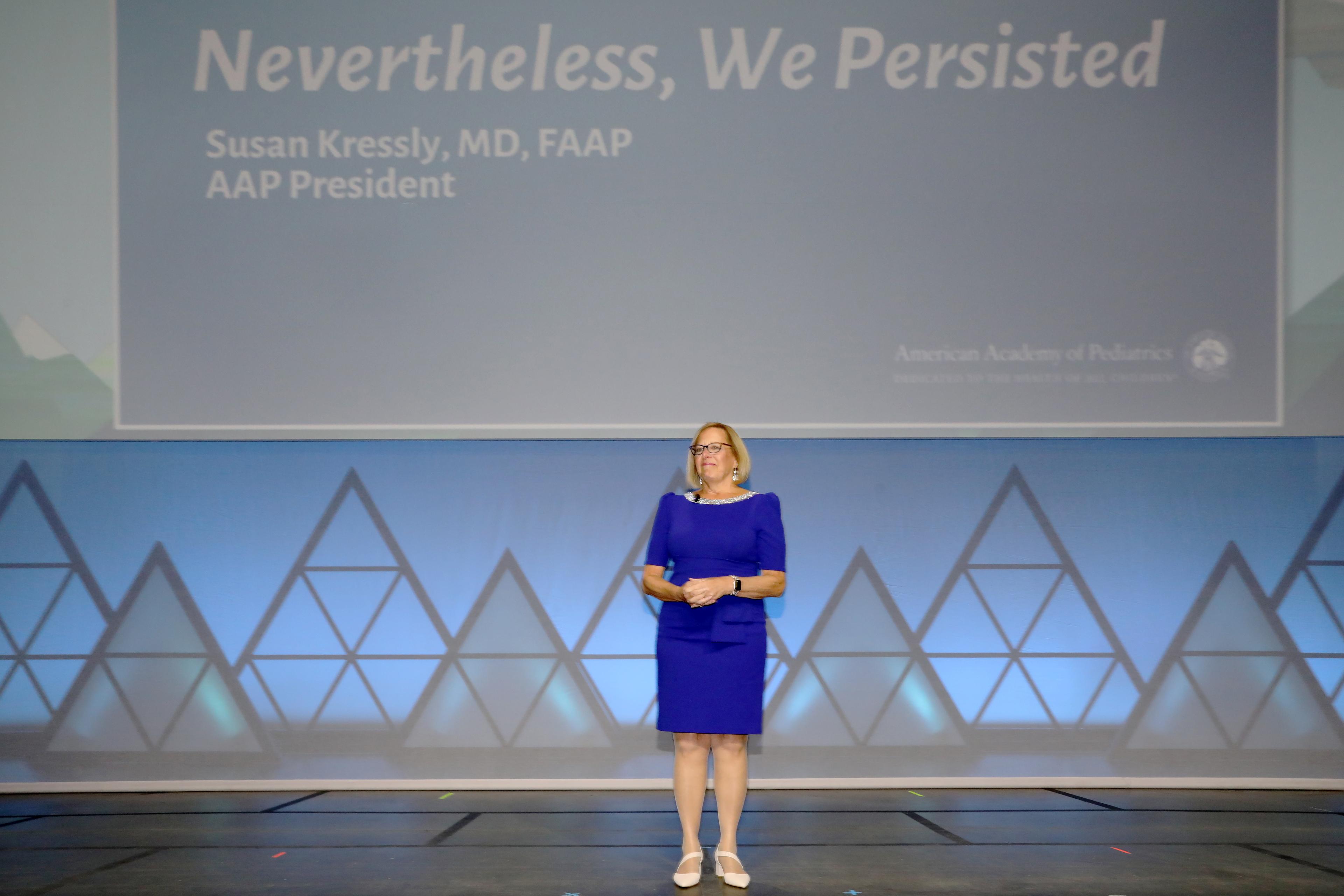 A woman in a blue dress stands in front of a large screen with words that say Nevertheless, we persisted