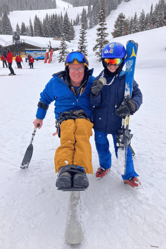 Man in a blue jacket and yellow pants is seated on a mono-ski in the snow facing the camera. A child also in blue with a blue helmet holding blue skis stands next to him.