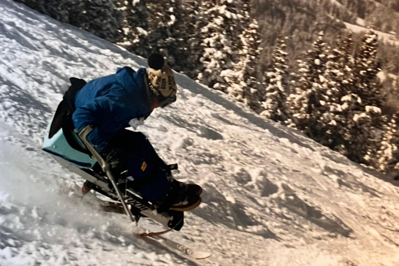 Image shows a snowy steeped mountain with a man in a blue jacket on a mono-ski heading down at a 45-degree angle.