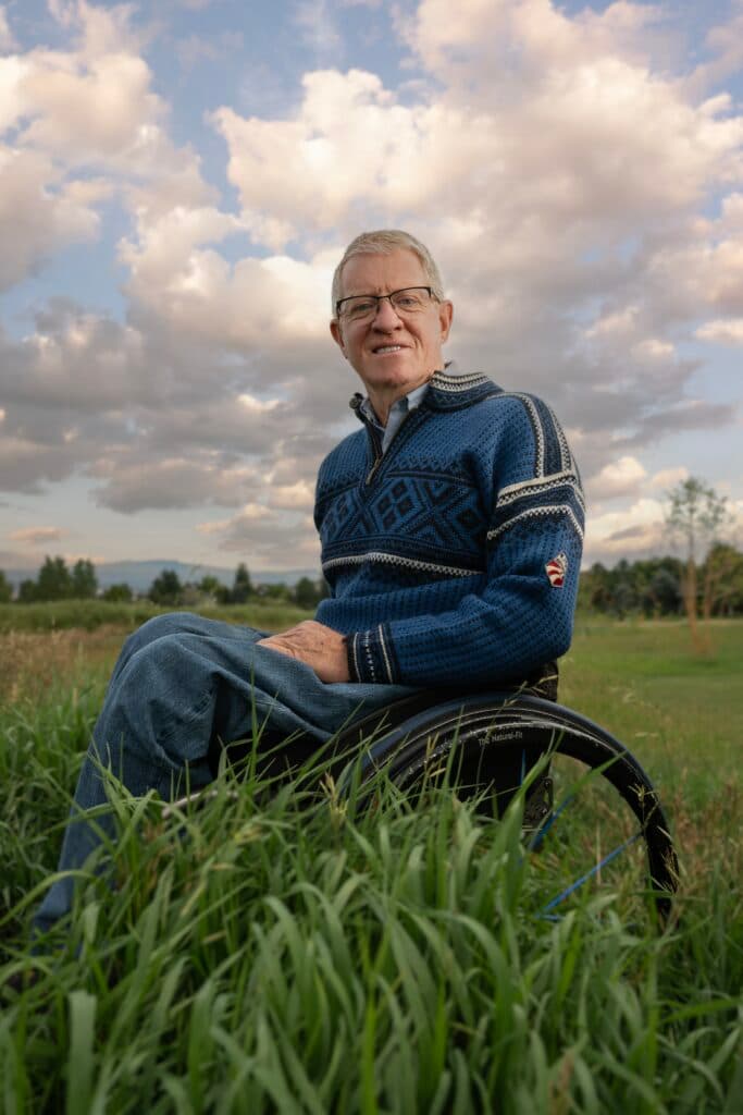 Man wearing blue is in a wheelchair sideways to the camera in a grass field and beneath a cloudy blue sky.
