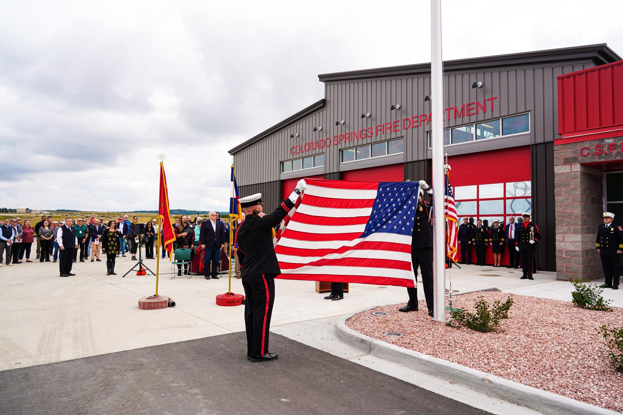people in uniform rase a flag in front of a firehouse