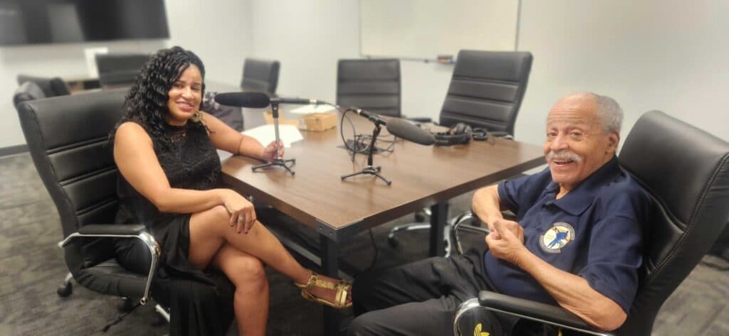 Woman in black dress is seated at a conference table next to a man in a dark blue shirt and black pants. There are black chairs around the table.
