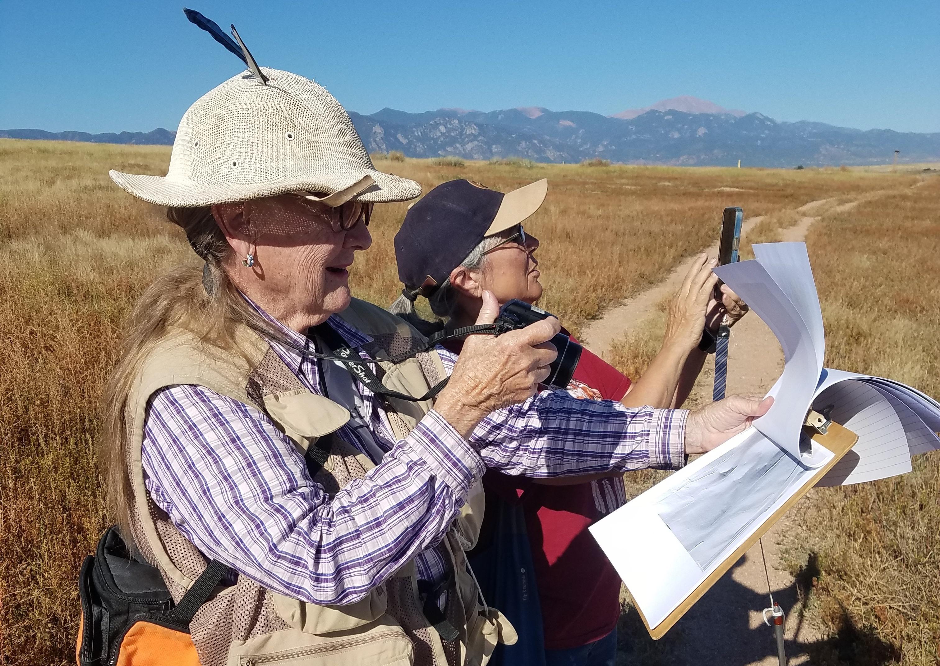 two people wearing outdoor gear and holding maps with the prairie and mountains in the background